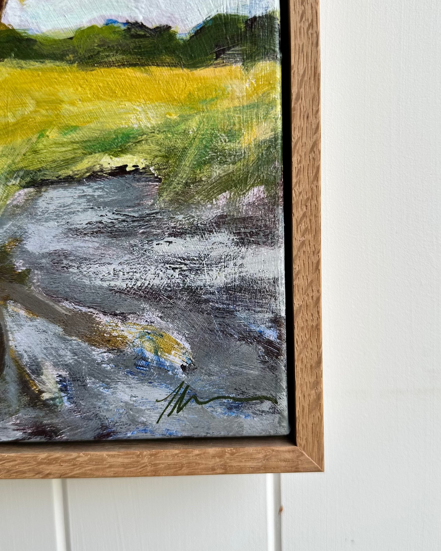 Framed painting of a landscape with a river and green fields on a white wall.