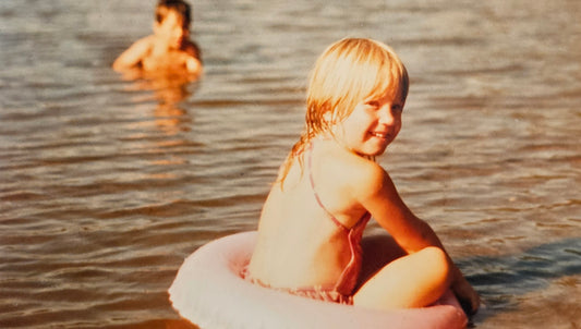 Terri as a little girl enjoying the morning sunshine, sitting in a pink floating ring on a river. In the background, her brother splashes in the water.