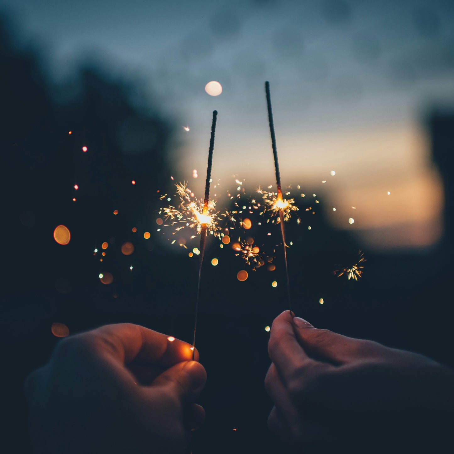 Two hands holding sparklers against a dark sky with a blurred background