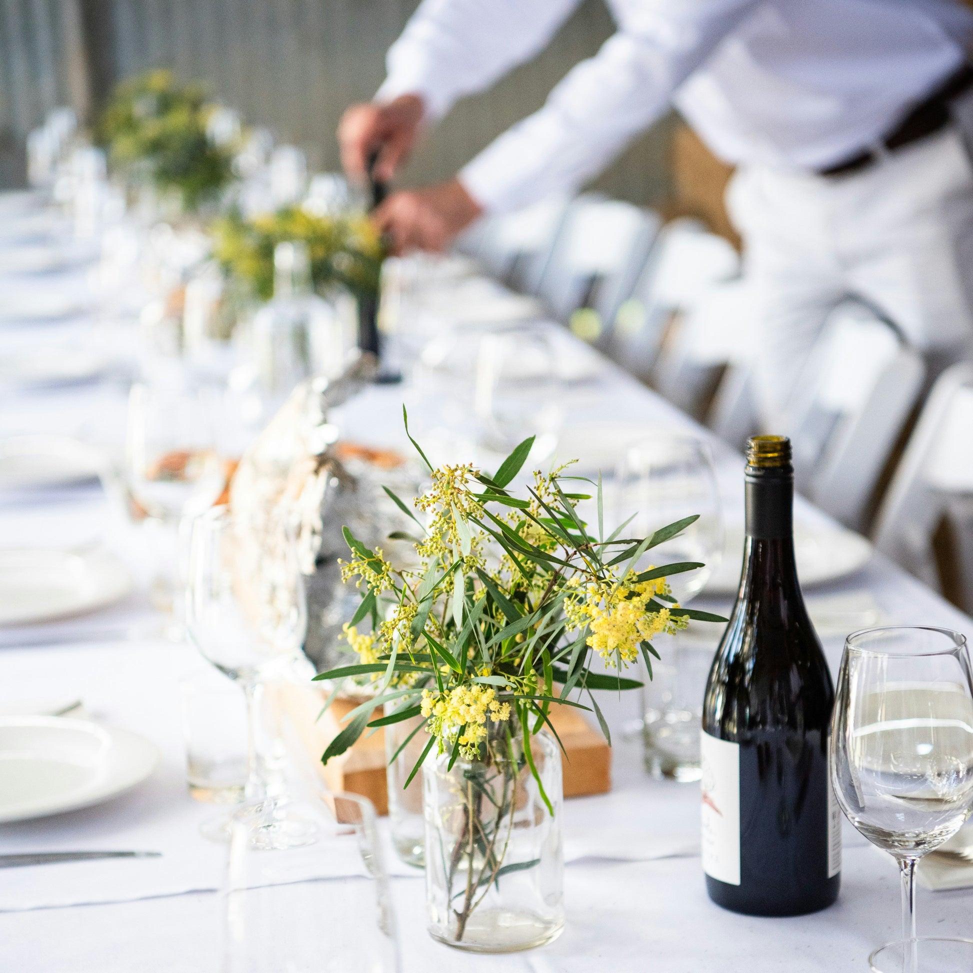 Long table set for a meal with white tablecloth, plates, glasses, and centerpieces.