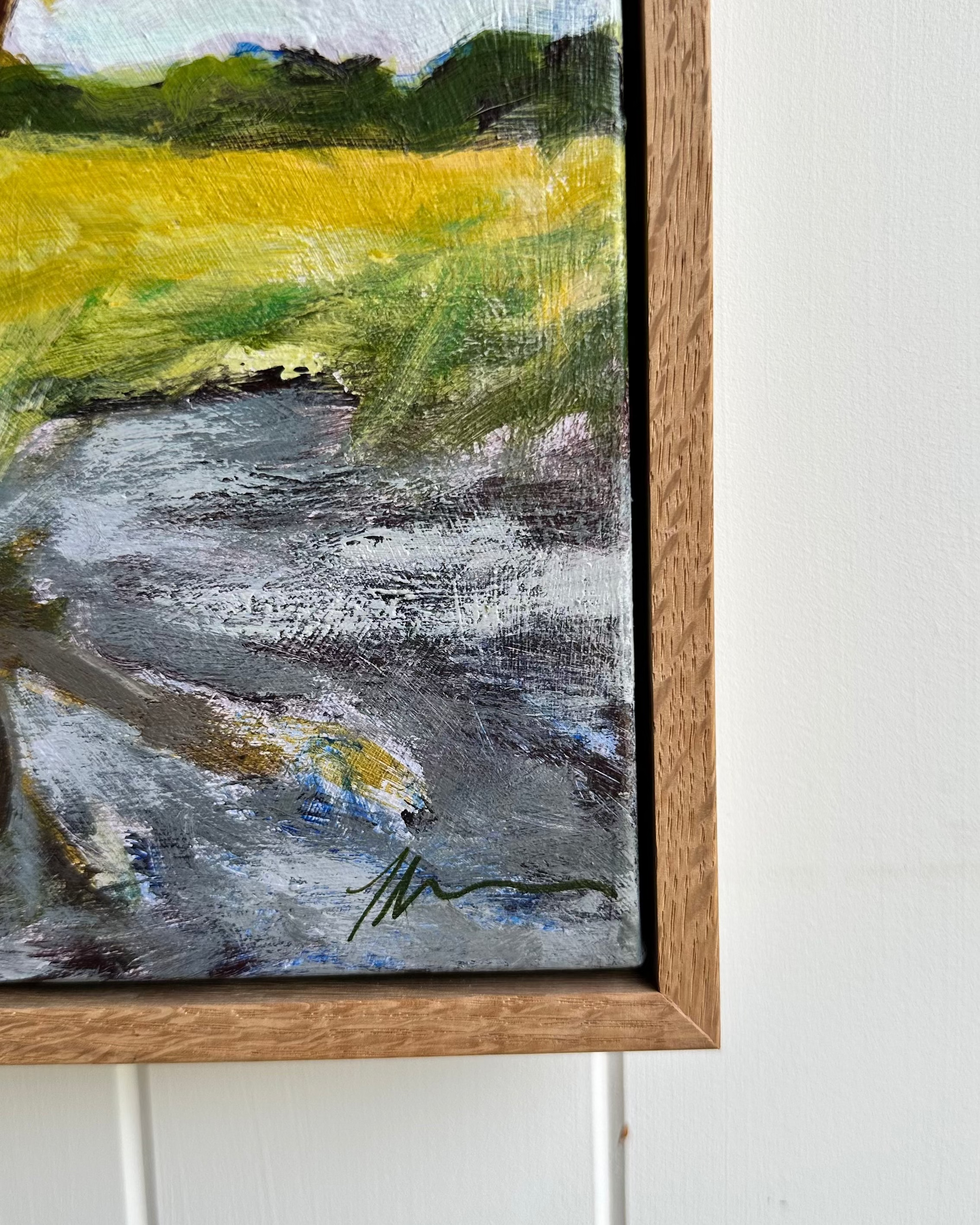 Framed painting of a landscape with a river and green fields on a white wall.